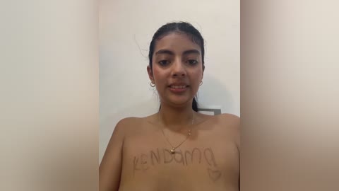 A video of a young, topless Latina woman with medium-brown skin and dark hair, standing in a narrow, beige-walled room. She wears hoop earrings and has a small tattoo on her left shoulder.