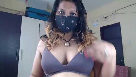 A video of a woman with medium skin tone, wearing a black face mask, gray bra, and a black necklace, standing indoors in a dimly lit room with closed cabinets and a doorway.