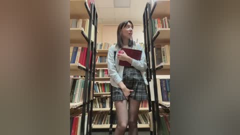 Video of a young woman with light skin, shoulder-length brown hair, wearing a grey sweater, plaid skirt, and sheer tights, standing in a library, holding a red book, surrounded by bookshelves.