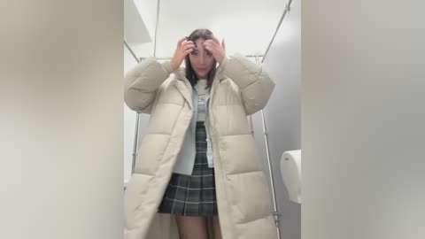 Video of a young woman in a plaid skirt, beige puffer coat, and grey sweater, covering her eyes with both hands in a public restroom stall.