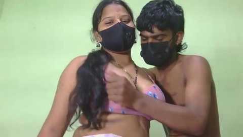 A video captures a South Asian couple, both masked, embracing; the woman, with long black hair, wears a pink floral bikini top, while the man, with dark hair and beard, is shirtless.