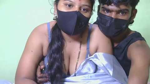 Video of two South Asian individuals with medium skin tones, wearing black face masks, black tank tops, and a silver robe, against a light green wall, taken indoors.