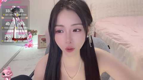 Video of a young Asian woman with long black hair, wearing a pearl necklace and a black top, in a pink bedroom.