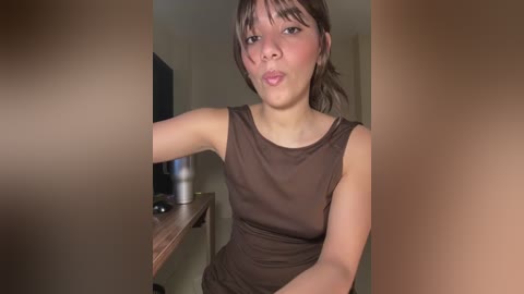 Video of a young Asian woman with light skin, brown eyes, and straight brown hair. She is wearing a sleeveless, form-fitting brown dress. The background shows a blurred, beige interior with a TV and a wooden table.