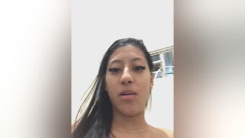 Video of a young, light-skinned woman with straight, dark hair, wearing a gold necklace, indoors against a white background, slightly blurred.