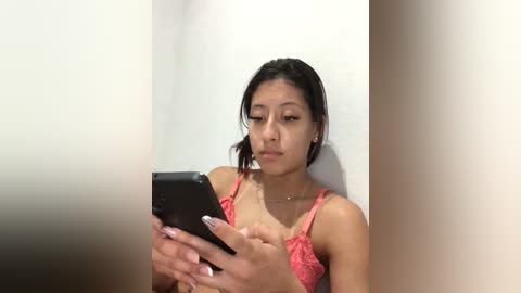 Video of a young Asian woman with straight black hair, wearing a pink lace bra, looking at her smartphone, in a simple, white-walled room.