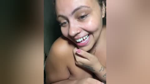 Video of a smiling, topless woman with light brown skin, dark hair, and a tattoo on her hand, in a dimly lit room.