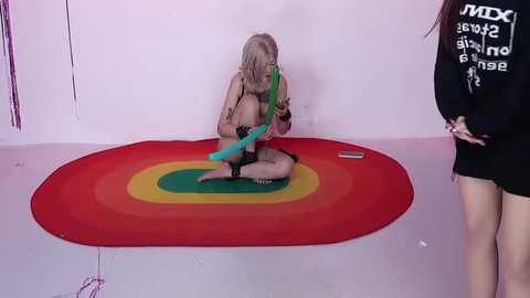 Video of a nude blonde woman with a blue dildo, kneeling on a rainbow mat in a minimalist room, facing a black-clad woman with a white t-shirt.