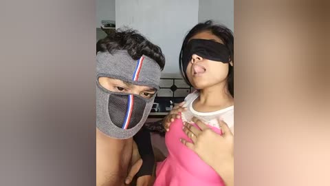 Video of a man in a gray mask with black stripes, and a young woman in a pink top, blindfolded, in a dimly lit room.