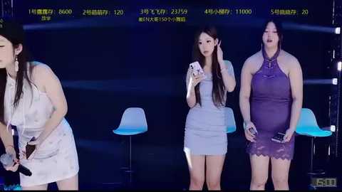 A video of three women in different dresses, white, light blue, and purple, standing in front of a blue-lit stage with a large screen displaying numbers.