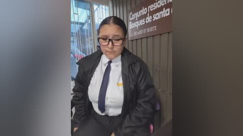 Video of a middle-aged Latina woman with medium skin tone, short black hair, glasses, and a black blazer over a white shirt and dark tie. She sits in front of a sign reading \"Capitan\u00eda de Puerto\".