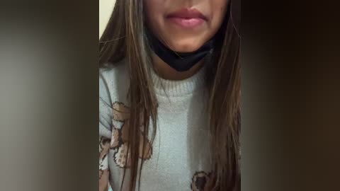 Video of a woman with long, straight brown hair wearing a white sweater with brown bear designs, a black blindfold, and pink lipstick, her face partially obscured by the blindfold.