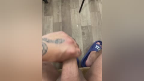 Video of a close-up of a person's hand with a tattoo on the wrist, holding a blue slipper, in a bathroom with a beige and brown tiled floor and a shower curtain.