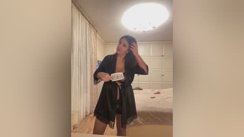 Video of a slender woman with long dark hair, wearing a black satin robe, brushing her hair in a bedroom with beige walls, wooden floor, and a bed with a beige blanket.
