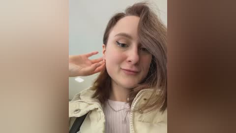 Video of a young woman with light skin and shoulder-length brown hair, smiling, wearing a beige puffer jacket, with a hand gently touching her ear.