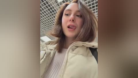 A video of a young woman with light skin and shoulder-length brown hair, wearing a beige puffer jacket over a white sweater. She is sticking her tongue out in a playful manner. Background shows a tiled ceiling.