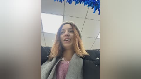 Video of a young woman with light skin and shoulder-length, straight, light brown hair, wearing a gray beanie and pink sweater, sitting on a black couch in a brightly lit room with white ceiling tiles and blue tinsel decorations.