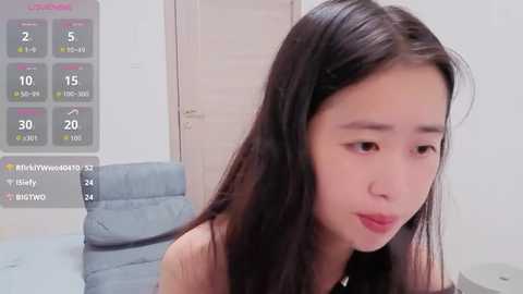 A video of an Asian woman with long black hair, light skin, and pink lipstick, sitting in a minimalist room with a blue sofa and calendar.