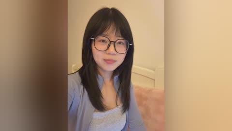 Video of an East Asian woman with long black hair, glasses, and a light blue top, standing in a beige room with a white bed and pink blanket.