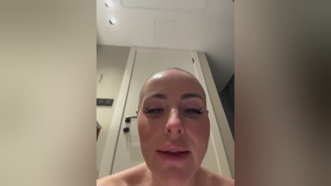 A video of a bald, light-skinned woman with heavy makeup, including black eyeliner and mascara, standing in a dimly lit hallway with white walls and a closed door.