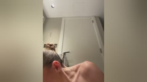 Video of a nude woman with blonde hair tied in a bun, seen from behind, standing in a bathroom with beige walls and a white door. A towel hangs on a hook.
