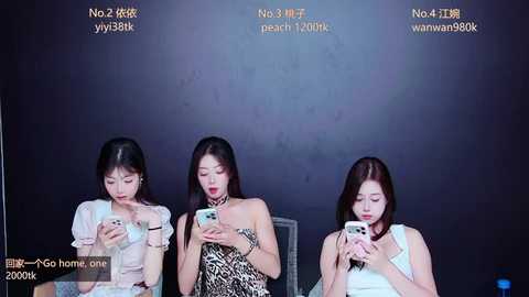 Video of three East Asian women, seated in a studio, using smartphones; one wearing a leopard-print dress, the others in casual attire, against a dark background.
