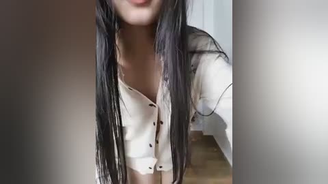 A video of a young Asian woman with long, straight black hair, wearing a white, button-up blouse, taken from a low angle. The background features a white door and wooden floor.
