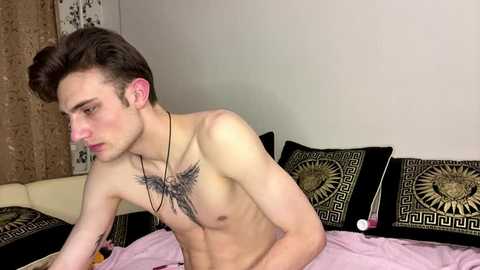 Video of a young, shirtless, lean Caucasian male with short, dark hair, prominent chest tattoo, sitting on a bed with black and gold geometric-patterned pillows, in a dimly lit room.