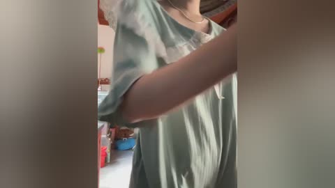 A video of a person in a mint-green blouse, seen from the shoulders up, with blurred background showing kitchen appliances and colorful containers.