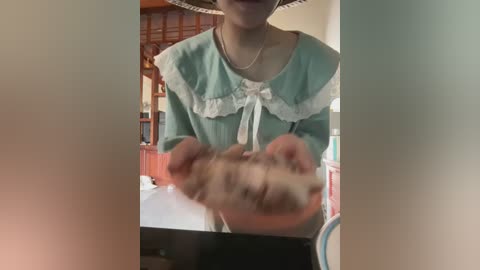 Video of a person in a green dress with white lace trim, holding a large, blurry, possibly fake, penis-shaped object. The background shows a wooden interior with a chair and shelves.