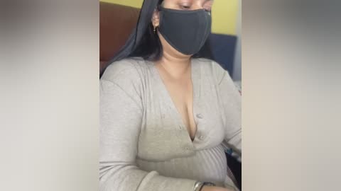 A video of a woman wearing a black face mask, a light gray ribbed V-neck top, and a silver watch, seated indoors with yellow and brown walls in the background.