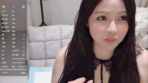 Video of an East Asian woman with long black hair, fair skin, and a delicate facial structure, wearing a black lace choker, sitting on a gray tufted bed. Background includes a bedside lamp and a white pillow.