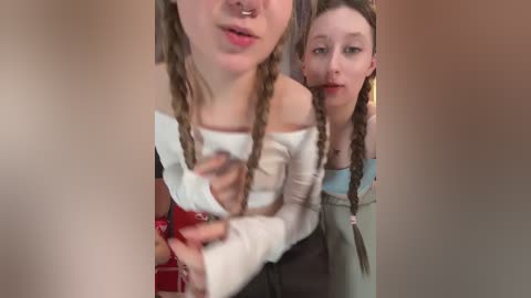 Video of two young women with long, braided hair. One wears a white off-shoulder top, the other a green sweater. They're smiling, indoors, with a blurred background.