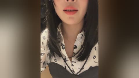Video of an Asian woman with straight black hair, wearing a white shirt with black floral patterns, and a black necklace.