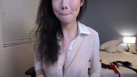 A video of an East Asian woman with long black hair, fair skin, and a white blouse, partially unbuttoned, smiling. Background includes a bed with white sheets, a lamp, and a window with closed blinds.
