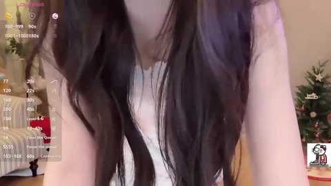 A close-up video of a woman with long, straight black hair wearing a white tank top, taken indoors with a Christmas tree and a bed in the background.