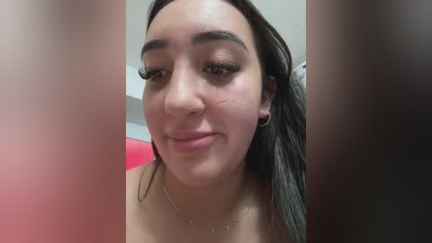 Video of a young woman with medium brown skin, long dark hair, and prominent eyebrows, wearing subtle makeup and a thin gold necklace. Background is blurred, likely indoor setting.