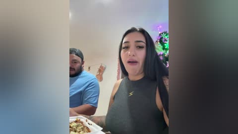 Video of a woman with long black hair and medium skin tone, wearing a sleeveless dark top, eating pizza, standing next to a man in a blue shirt and cap, in a brightly lit room with festive decorations.