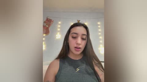 Video of a young woman with long dark hair, wearing a black sleeveless top with a lightning bolt design, in a softly lit room with string lights and a Christmas ornament.