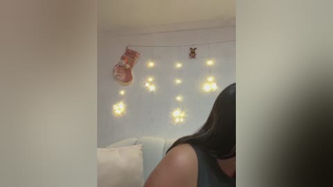 Video of a woman with long, dark hair, wearing a black sleeveless top, standing in a softly lit room adorned with white walls, a white couch, and a string of warm, glowing fairy lights.