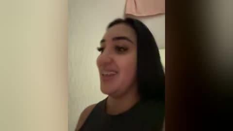 Video of a young woman with straight black hair, fair skin, and a dark sleeveless top, smiling slightly. Background features a beige wall with a pink towel hanging above.