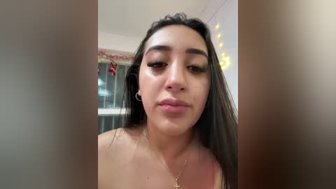 Video of a young woman with medium skin tone, long dark hair, and full lips, wearing a delicate gold necklace. She's indoors, near a window with a red ornament.