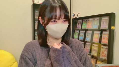 Video of an East Asian woman with straight black hair and fair skin, wearing a beige mask, purple sweater, and yellow clothing. She's indoors, near a colorful shelf of books.