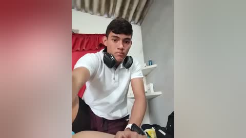 Video of a young, fit, light-skinned male with short dark hair, wearing a white polo and maroon shorts, sitting on a bed with red curtains, white shelves, and a black bag in the background.