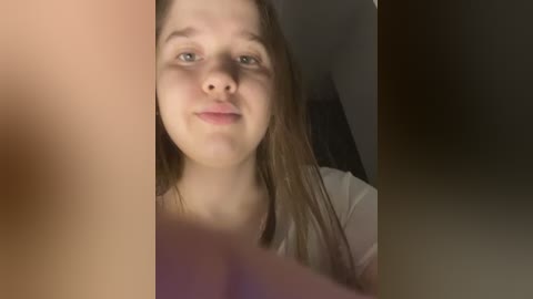 Video of a young Caucasian woman with straight brown hair and fair skin, smiling slightly. She is wearing a white top. The background is dimly lit with shadows.