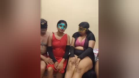 Video of three people in Halloween costumes: a woman in a red dress, a man in a black shirt, and a woman in a black dress and mask, sitting on a bench.