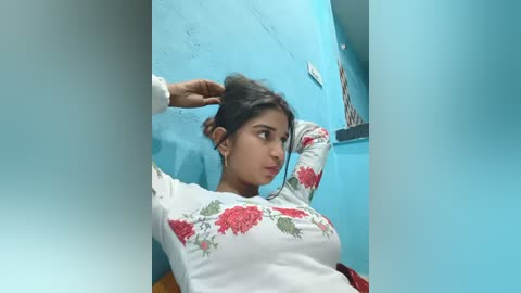 Video of a South Asian woman with medium skin tone and long black hair, wearing a white long-sleeved shirt with red floral embroidery, standing in a small, blue-painted room with a white wall and a framed photo.