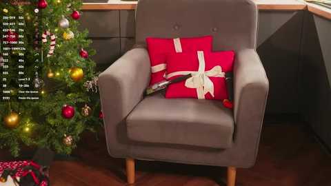 Video of a modern living room with a gray armchair featuring a red pillow and wrapped gift, next to a decorated Christmas tree.
