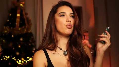 Video of a woman with long, wavy brown hair, wearing a black top, blowing a kiss while holding a green drink. Background features a lit Christmas tree with gold ornaments.