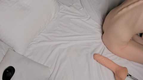 Video of a fair-skinned woman lying on a white bed, partially covered by a white sheet, with a large, light-skinned dildo resting on her stomach, and a black remote control nearby.